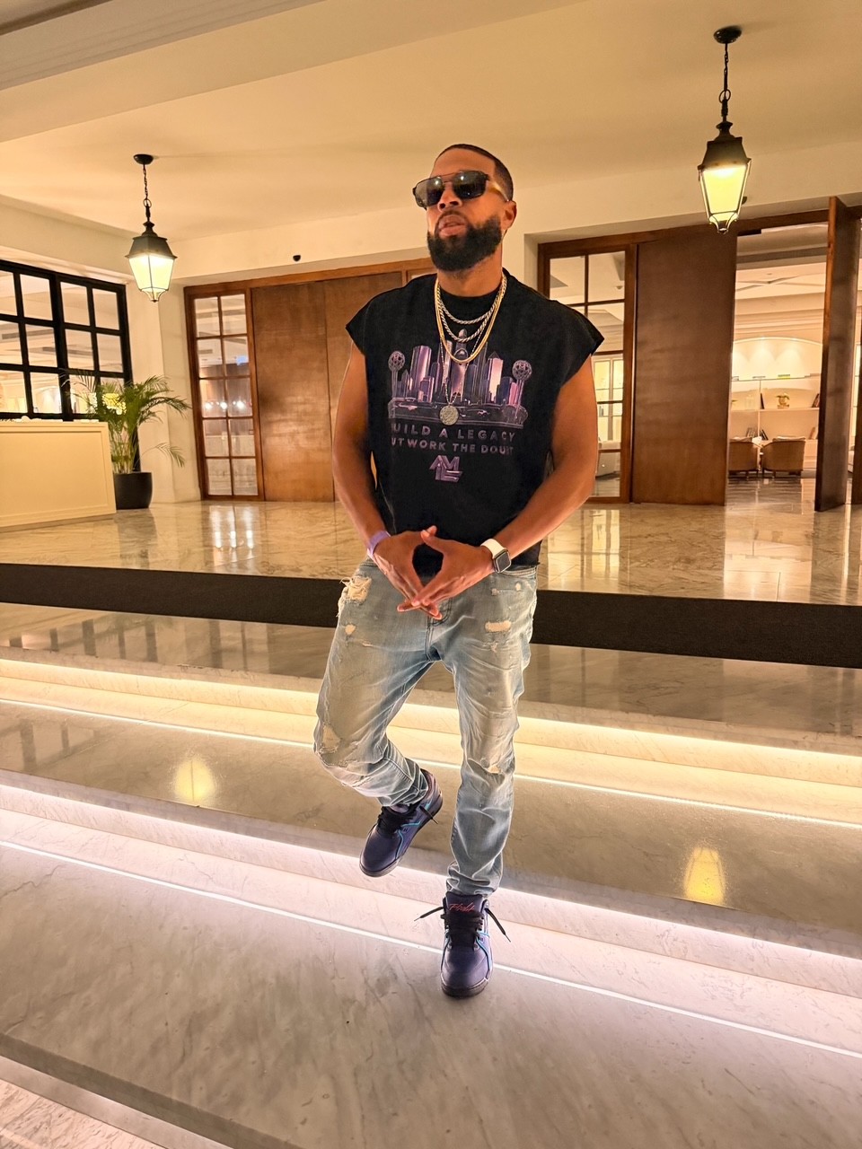 DAVĒ wearing sunglasses, a black graphic shirt and jeans on illuminated lobby steps.