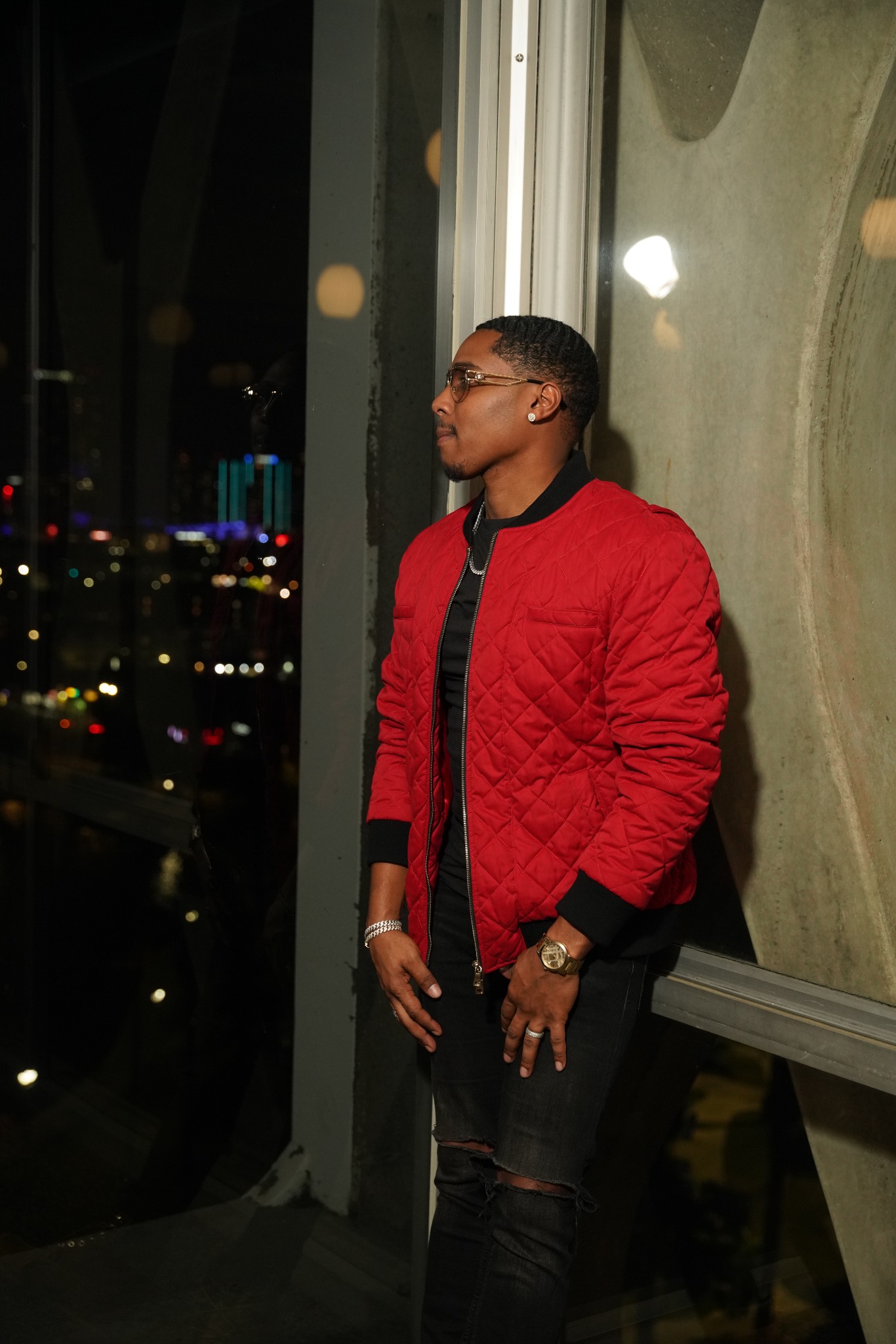 DevAMIL in a red jacket and glasses beside a window overlooking city lights.