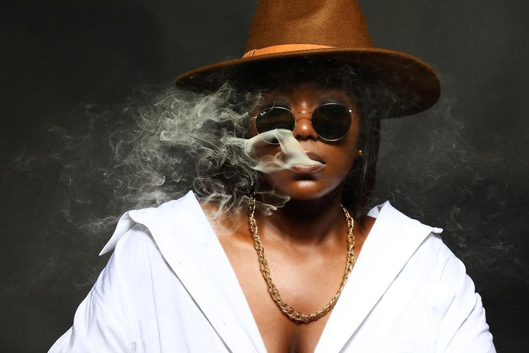 Diamonique Jackson wearing a brown wide-brim hat, round sunglasses, a white shirt and gold chain against a dark backdrop.
