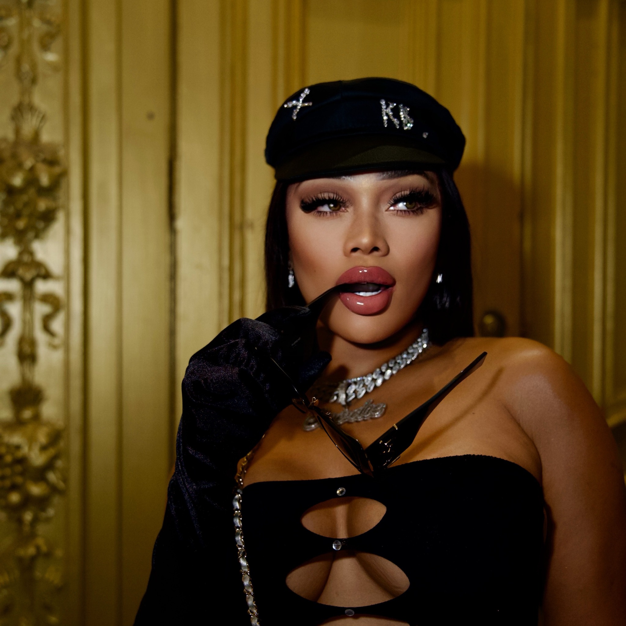 Gwan Goodie in a black outfit and cap against a gold backdrop.