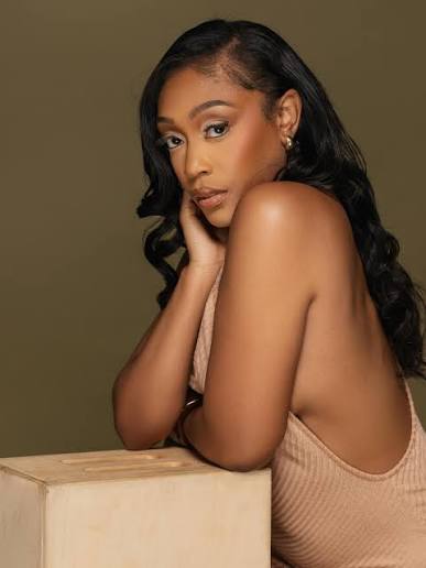JaQuandice leaning on a wooden pedestal against an olive backdrop.
