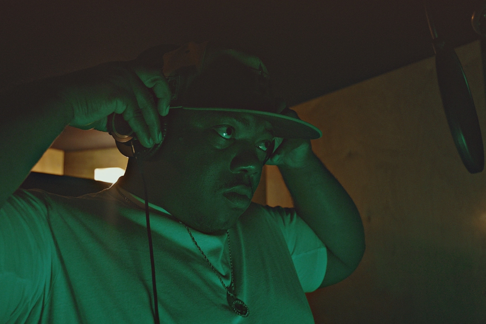 Vicious J wearing headphones beside a studio microphone under green and amber lighting.